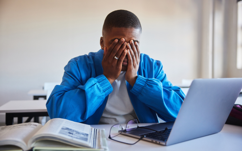 Student with his hands on his face at a desk with a computer, glasses, and book