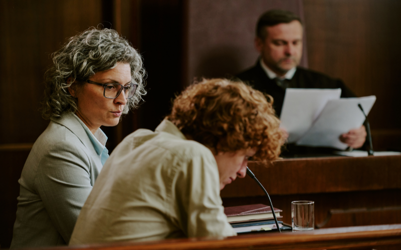 In a courthouse, female lawyer next to a teen boy at a table with a judge in the background