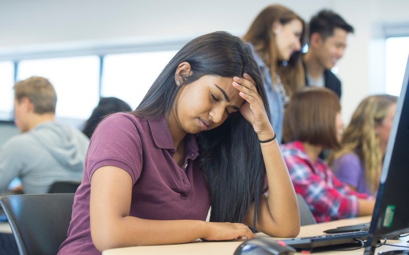 Stressed college student holding her head in her hand while sitting in front of a computer, other students talking in a group in the background