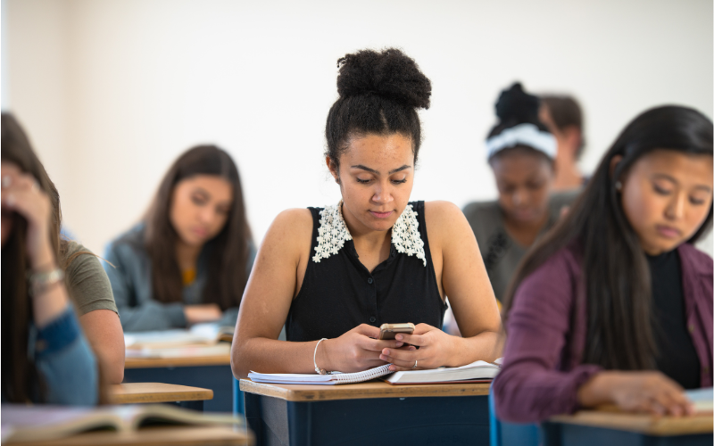 Student looking at a cell phone while sitting at a desk surrounded by other students in a classroom
