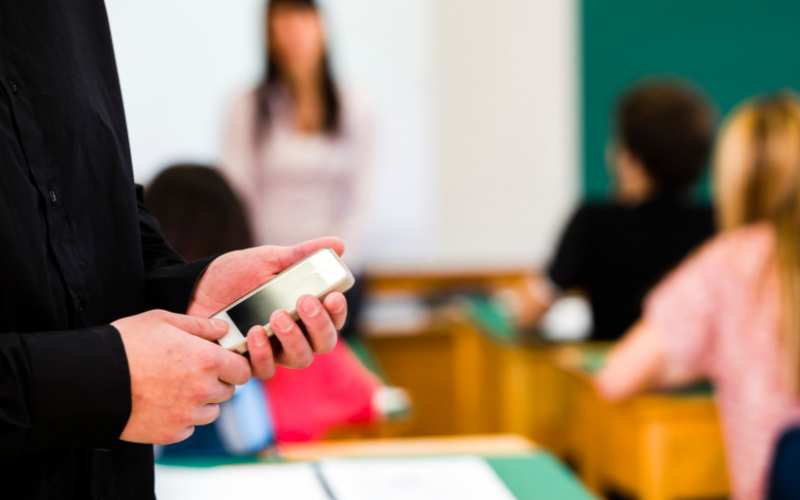 Person looking at cell phone in forefront, blurred students and teacher in classroom in background