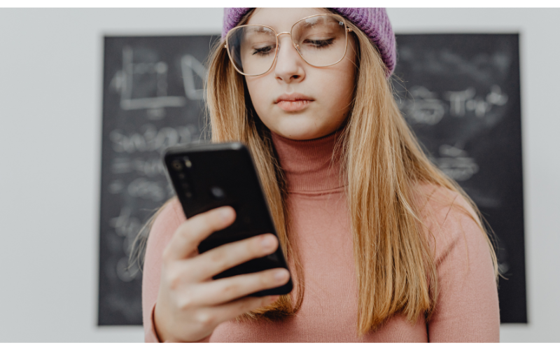 Teen in glasses and hat holding a cell phone with blackboard behind her