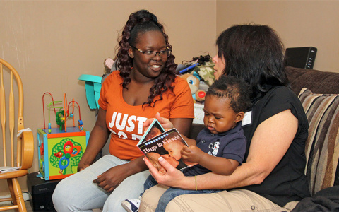 A healthcare worker visiting a young mother. The infant is reading a pictured book in his mom's lap.
